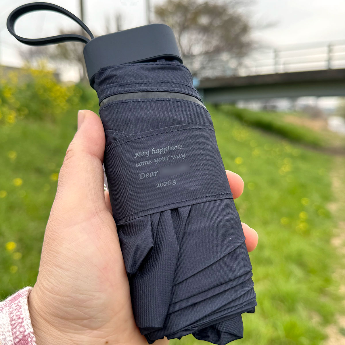 春の菜の花が咲く小川沿いで撮影した、メッセージ刻印入りの名入れ折りたたみ傘。温かな応援メッセージが添えられた世界にひとつの雨具