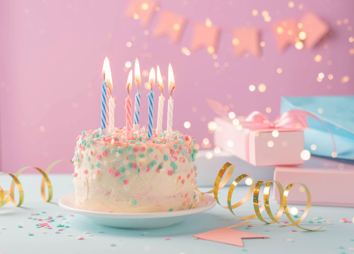 キャンドルが灯るバースデーケーキとピンクの背景に飾られたギフトボックス。大切な人の誕生日を祝う華やかなパーティーシーン。