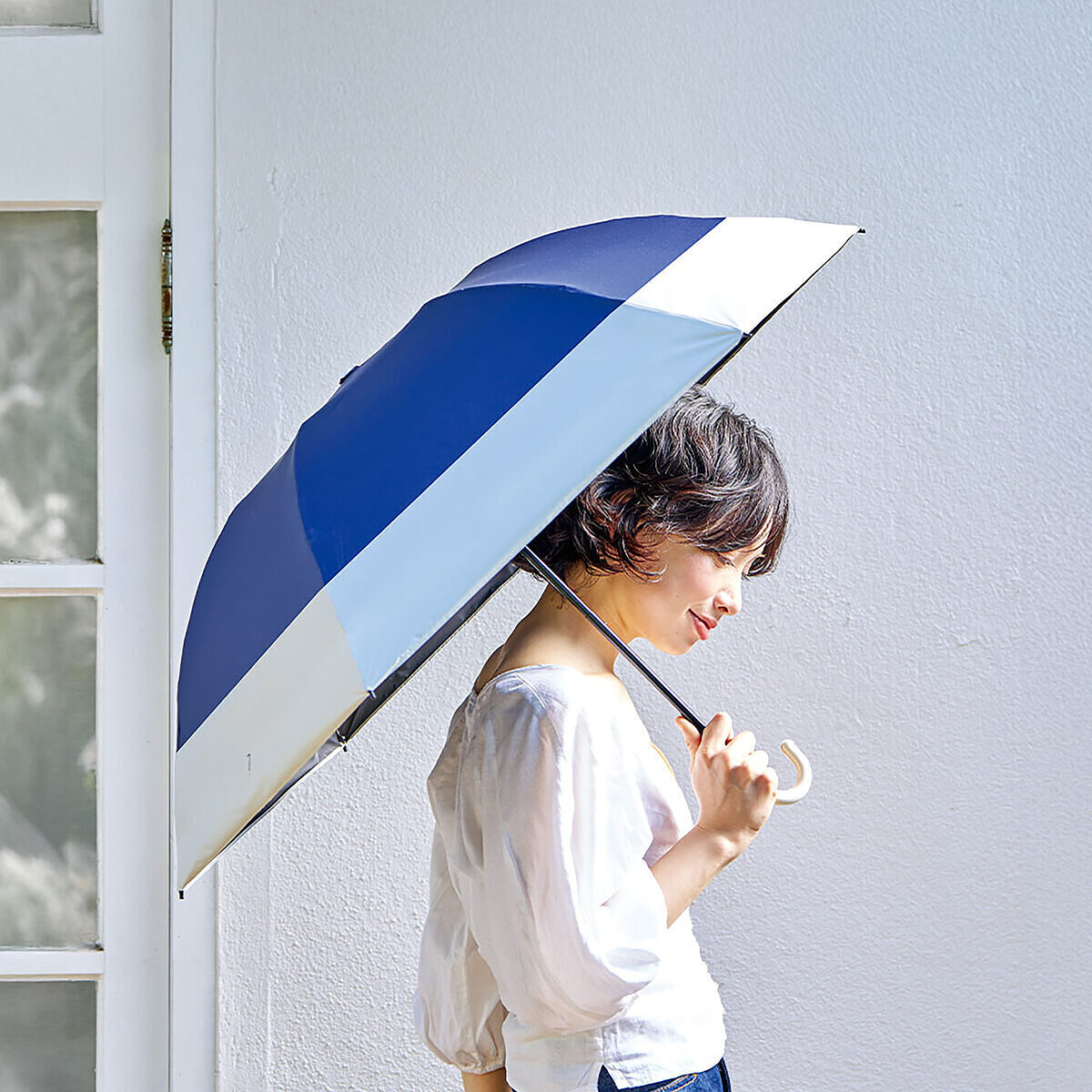 【着用イメージ】晴雨兼用の「ツートーン折りたたみ傘」を開いてさしている女性の写真。梅雨の曇り空でもファッションを引き立てるお洒落な配色デザイン